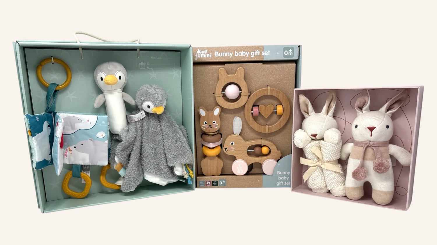 Three wooden baby gift sets displayed side by side on a light beige background: a penguin and duck soft toy set in a mint box, a wooden bunny teething ring set in a brown box, and a pair of plush bunny toys in a pink box.