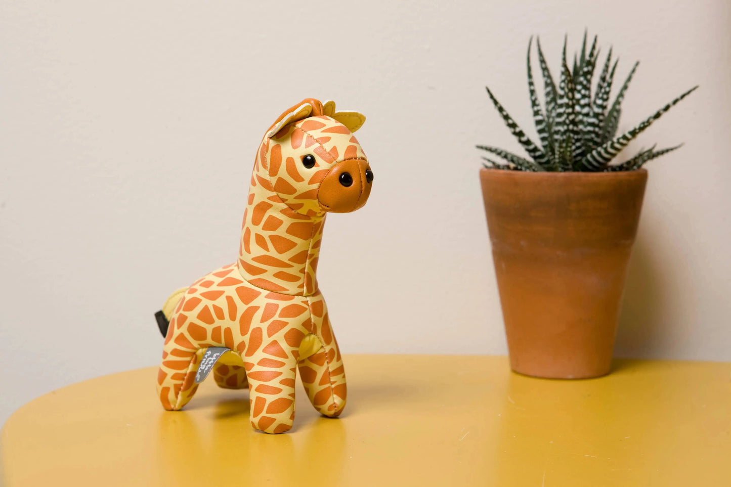 Plush giraffe toy on a yellow surface with a potted plant in the background