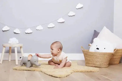 Baby playing with a toy elephant on a rug in a room with a basket and cloud decorations.